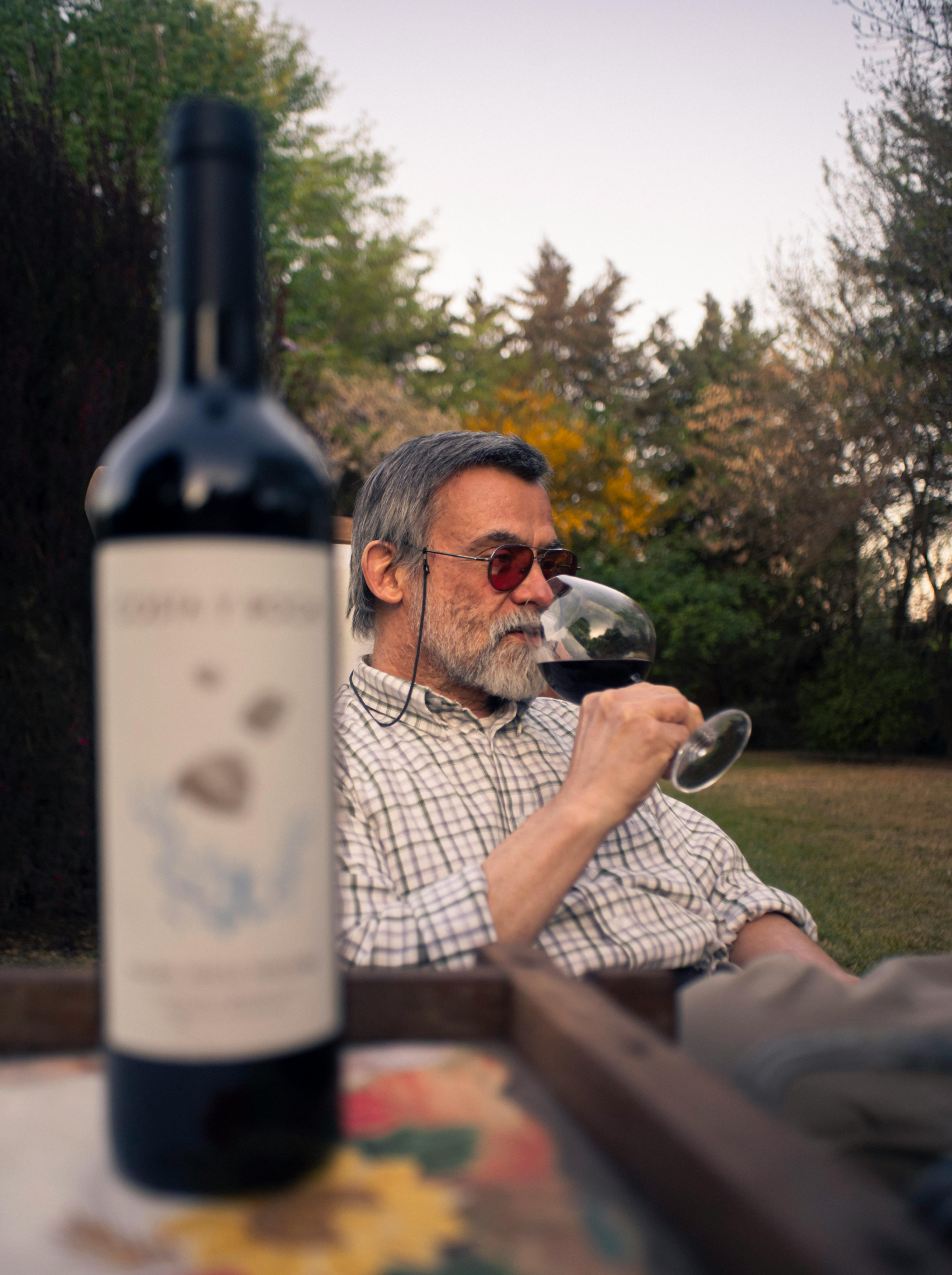 Man sitting in chair siping on a glass of red wine in a field with trees and a bottle of wine in the fore ground on a table blurred